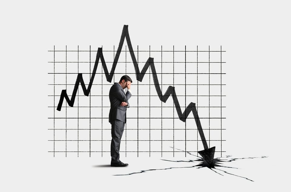 Businessman in a gray suit standing with his hand on his forehead, facing a jagged line chart on graph paper that plunges downward and cracks through the floor where its arrow lands.